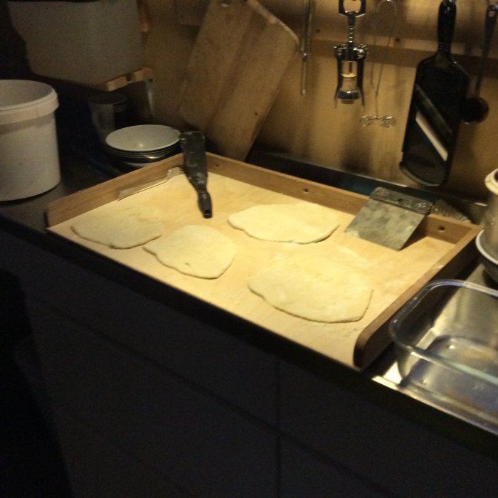 Fyra degklumpar för spisbröd på en upphöjd träyta i ett kök med köksredskap hängande ovanför.