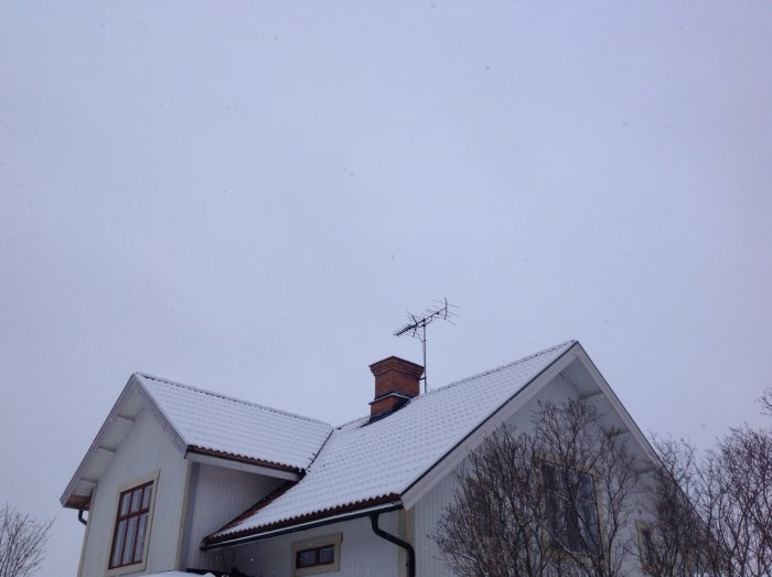 Hus med snötäckt sadeltak och skorsten, antennen på taket, omgiven av disig himmel.