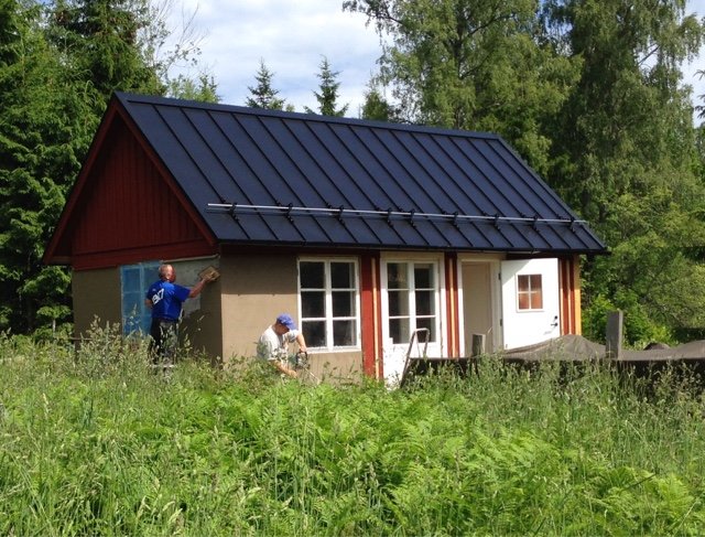 Två personer som arbetar på att putsa en mur på en liten stuga med röda fönsterfoder och svart tak.