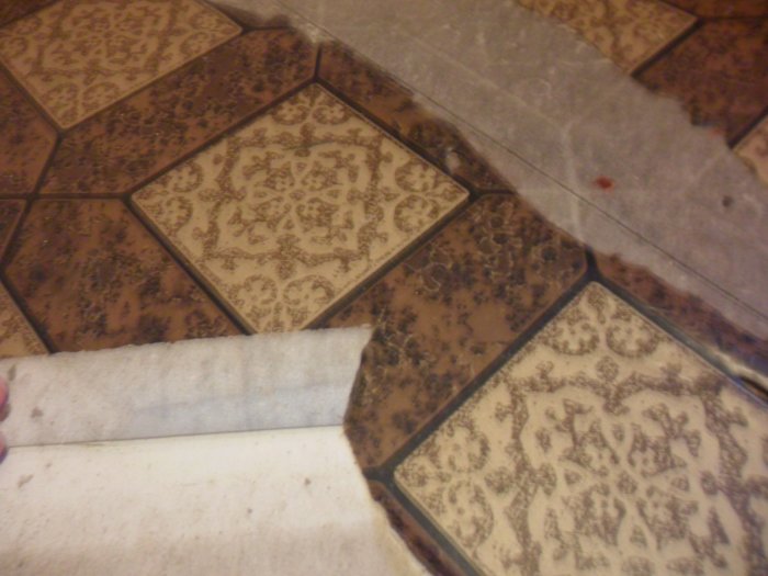 Mönstrade, bruna och beige kakelplattor. Vit kant. Olika material möts på sliten yta. Nedslitet golv.