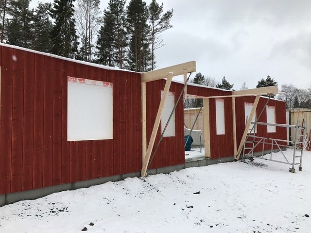 Ny uppsatt röd träpanel på husets fasad med utbyggnad för farstukvist under konstruktion, omgiven av snö.