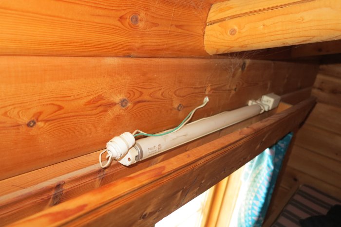 En lampamontering under en väggfast säng med synliga kablar och spindelnät.