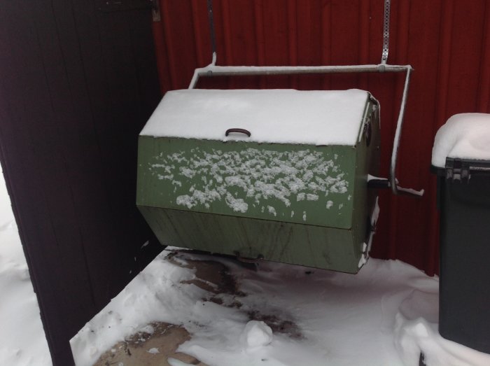 Grön kompostbehållare täckt med snö bredvid en soptunna ute i vinterväder.