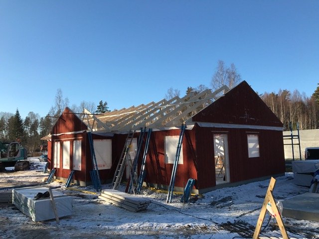 Takstolar på ett hus under konstruktion med flera dörröppningar och byggmaterial runtomkring.