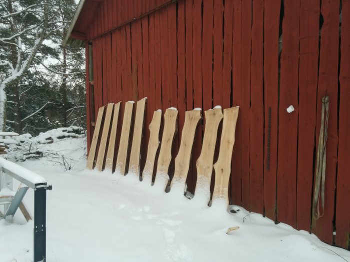 Stora träplankor av ask uppradade mot en röd stuga i snöig omgivning.