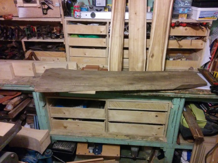 Träplanka på slitet verktygsbord i verkstad med snickeriverktyg i bakgrunden.