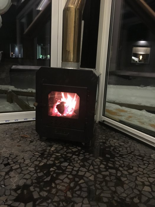 Vedeldad kamin med brinnande eld inne i ett rum, reflektion i fönstret och snö utanför.