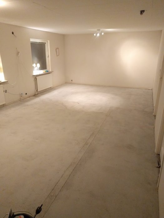 Avjämnat betonggolv i ett upplyst, tomt rum under renovering med synliga elkablar och en fönster.