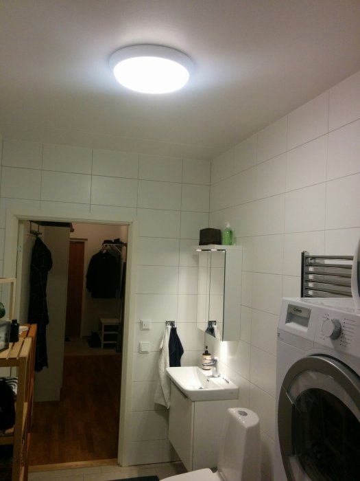 Badrum med starkt takljus, spegelskåp, tvättmaskin, handfat och toalett.