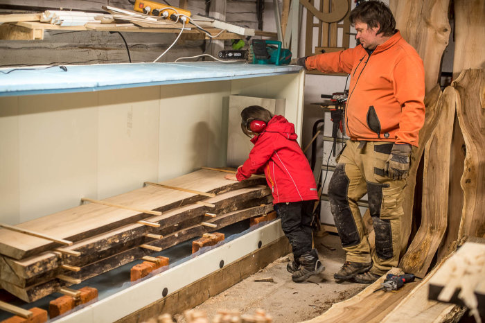 Barn hjälper vuxen att stapla träplankor i ett verkstadsutrymme.