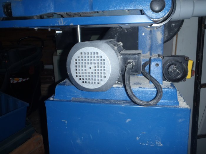 En nymonterad motor på ett blått band/skivputsverktyg med provisorisk plywoodremskiva, kablar, och motorbroms i ett verkstadsutrymme.