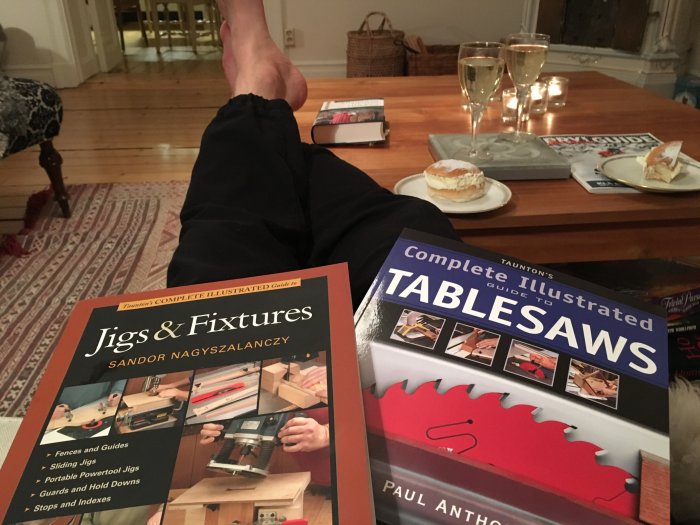 Relaxing evening with woodworking books, champagne, and a pastry on a table, person's legs reclined on a sofa.