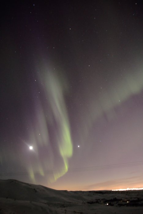 Norrsken över ett snölandskap på Island med stjärnor och en ljus stad i bakgrunden.