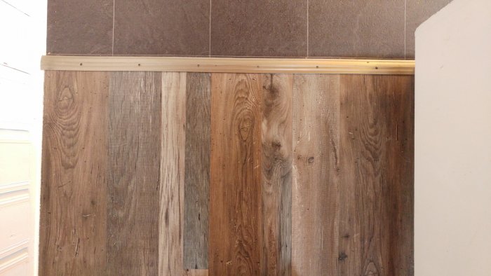 Träpanel och bruna kakelplattor ovanför en golvlist i ett rum.