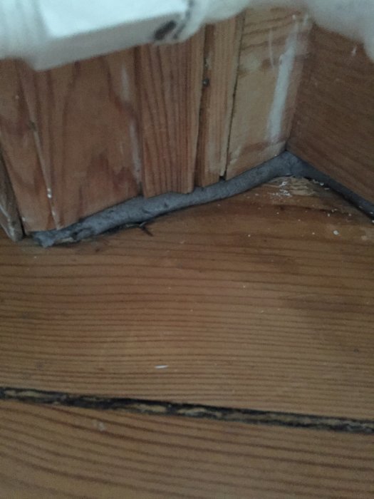 Gammalt trägolv som möter en vit vägg med en liten öppning där listen inte helt täcker gapet.