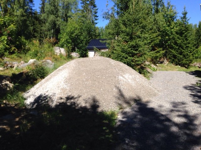 En stor hög av grus i en trädgård med gröna träd och en byggnad i bakgrunden, vilket indikerar ett pågående projekt.