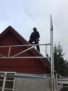 Person på byggställning bredvid rött hus med arbetskläder och hjälm, inget synligt arbete utförs.