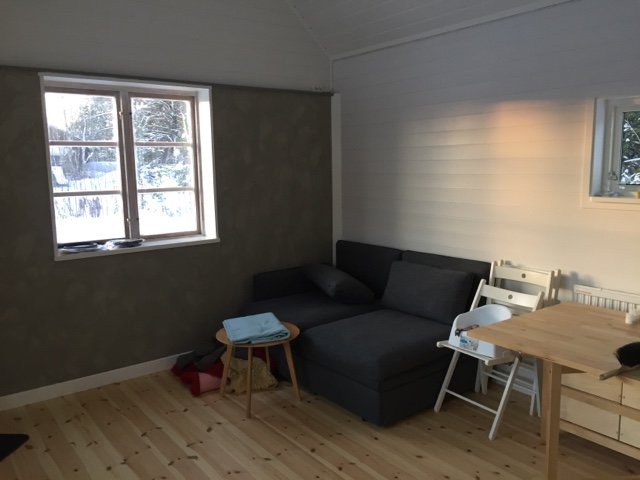 Inredning av lillstuga med bäddsoffa, matbord och stol, utsikt genom fönster mot snölandskap.