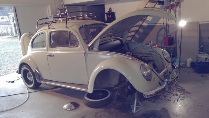 Vit Volkswagen Typ 1 ('Bubbla') på garagegolv med öppen motorlucka för underhåll.