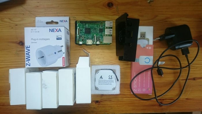 Hemautomatiseringskomponenter utspridda på ett träbord, inklusive Raspberry Pi, Z-wave USB stick, dimmer och förpackningar.