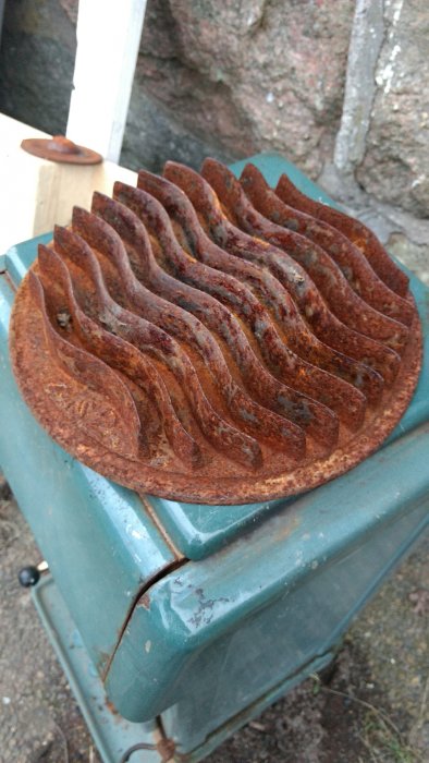 Rostig rund metallplatta med vågiga flänsar, ligger på en grön behållare utomhus.