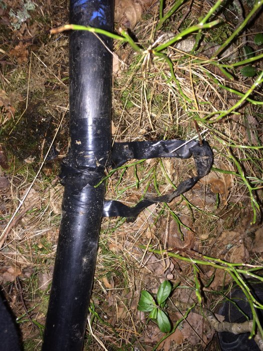 En svart plaströr med gammal eltejp som har gått sönder, vilket exponerar en skarv omgiven av gräs och löv.