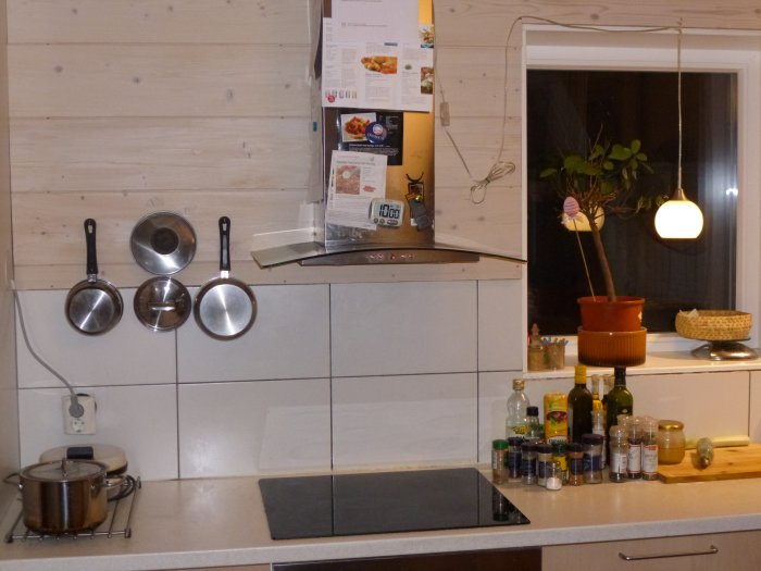 Ett kök med trävägg, vita kakelplattor, stekepannor upphängda, kryddor och oljor på bänk.