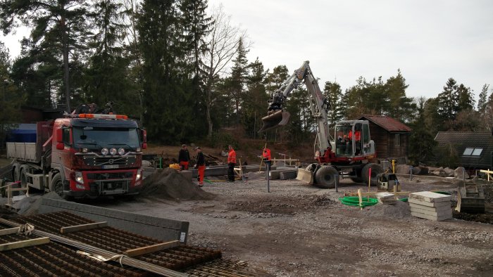 Byggarbetsplats med arbetare, avloppsrör och balkar synliga, lastbil och grävmaskin i arbete.