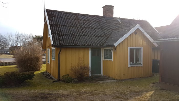 Ett gult trähus med svart tak och grön dörr mitt under renovering, omgivet av en trädgård.