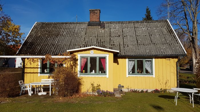 Ett gult hus med förfallet svart tak och röda gardiner synligt i renoveringsbehov under en klar himmel.