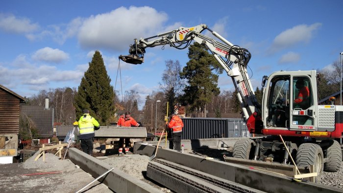 Arbetsplats med en grävmaskin som lyfter balkar och arbetare som granskar byggarbetet.