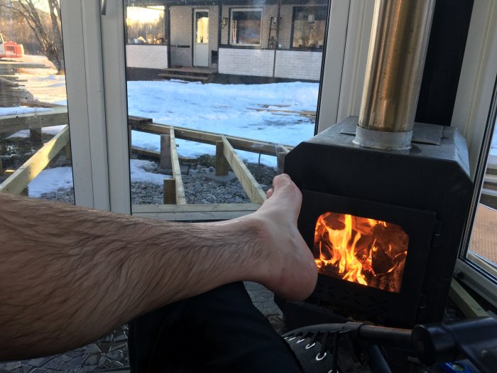 Person värmer sig vid en kamin med öppen eld inomhus, snötäckt uteplats synlig genom fönster.