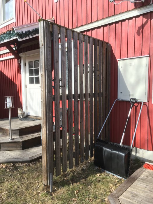 Ett cirka 2,4 meter högt trästaket framför en röd stuga, gråa plankor med mellanrum och snöstänger stödda mot det.