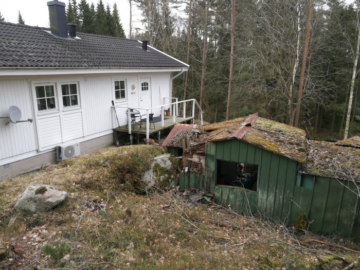 Ett gammalt sommarhus med slitet och mossaöverväxt tak nära ett gästhus och luftvärmepump.