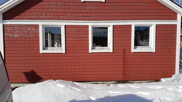 Röd träfasad med tre vita fönster på en byggnad, snö på marken framför och ojämn sill synlig längs husgrunden.