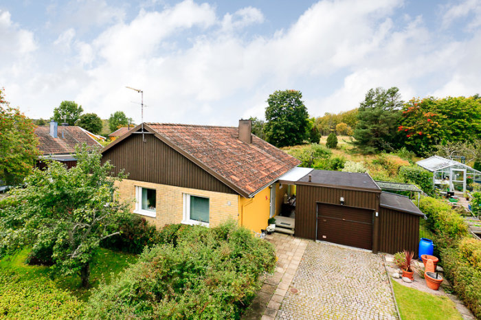 Ett hus och garage med tillbyggnad, trädgård i förgrunden, mäklarbild med miljö.