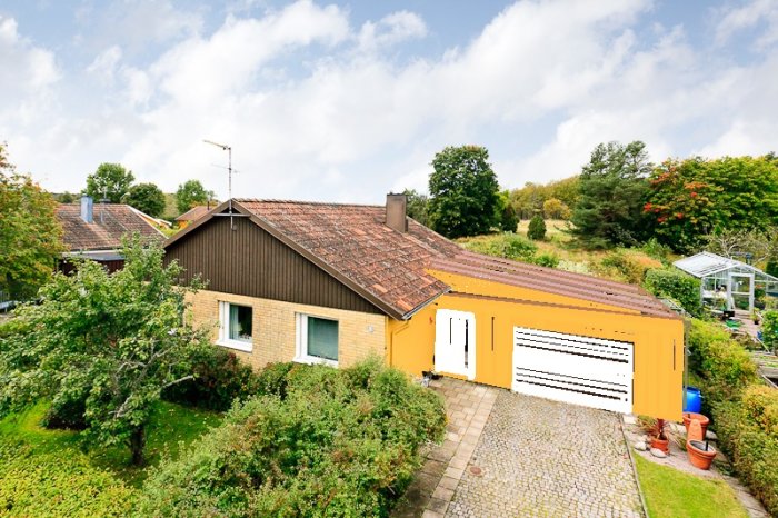 Vy över en enplansvilla med anslutande oisolerat garage och trädgård.