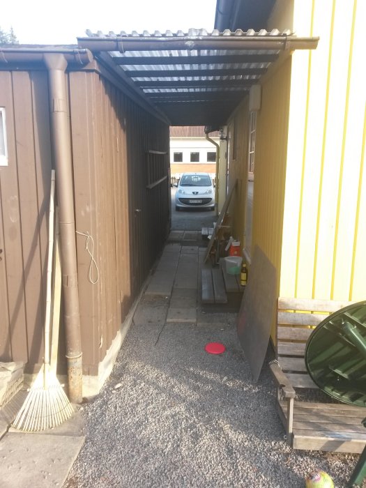 Smal passage mellan en huskropp och ett brunt garage med skjul och grusgolv som leder till en bilparkering.