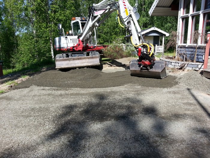 En grävmaskin i arbete med att utjämna grus och stenmjöl på en blivande uteplats vid ett rött hus.