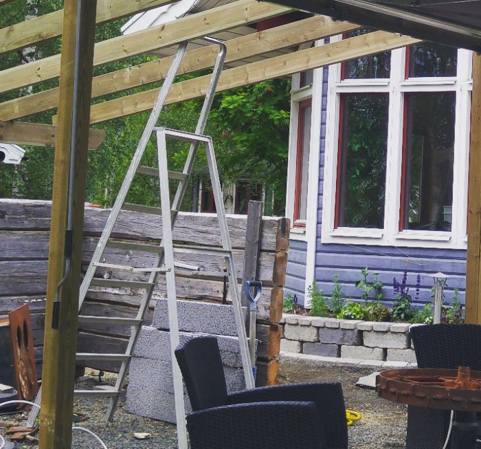 Byggprojekt med träkonstruktion och gammal järnspis, stockar från hus vid pergola under uppbyggnad.