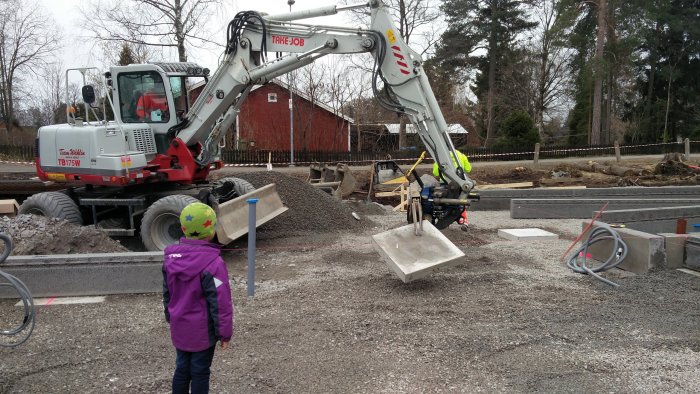 Grävmaskin och arbetare som placerar gråa trottoarkantstenar medan ett barn observerar byggarbetsplatsen.