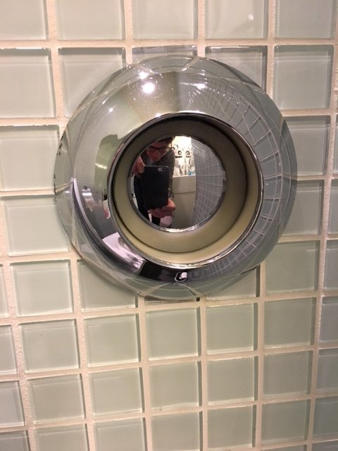 Rund spolknapp på en vägghängd toalett med spegelreflektion och kakelvägg i bakgrunden.