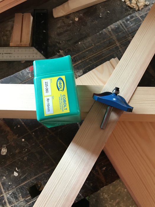 Fräsprofil på träplanka bredvid ett nytt frässtål för S-profil i dess förpackning, arbetsområde med verktyg i bakgrunden.
