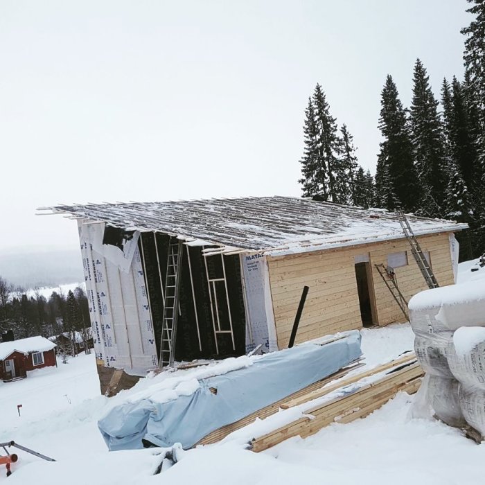 Underkonstruktion av tak på trähus i snöigt landskap med isoleringsmaterial och byggmaterial synligt.