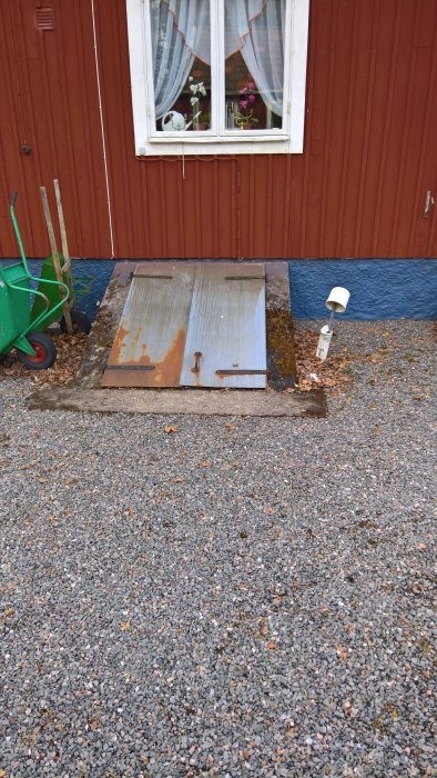 Tung dubbellucka i metall till källare framför röd trävägg och fönster, med hjulburen och mur vid öppningen.