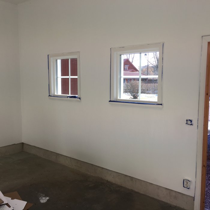 Ett rum med betonggolv som övergår i en vit vägg med två fönster, synligt mellanrum nära golvet där klickgolv ska läggas.