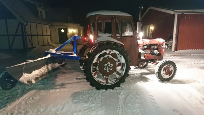 En röd traktor med plogblad framför justerar snö på gårdsplan i mörkret.