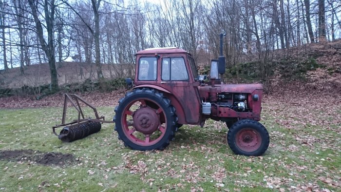 Röd traktor med ringvält på gräsmatta med löv och mullvadshögar i bakgrunden.