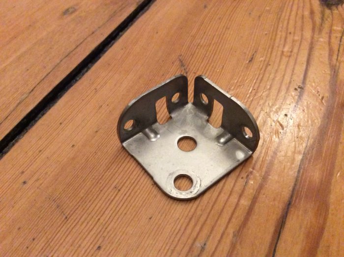 Metallfäste med fyra skruvhål på ett trägolv.