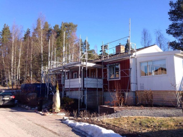 Hus med ställningar för takbyte, svarta betongpannor synliga, omgivet av snöfläckad trädgård.